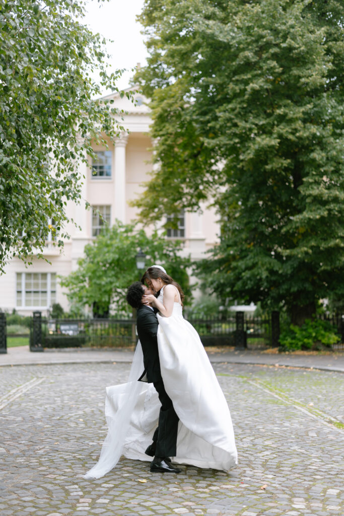 A groom in a black suit lifts and kisses a bride in a white wedding dress on a cobblestone path, with greenery and a large white building in the background.