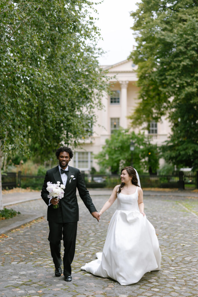 A bride in a white dress and a groom in a black tuxedo walk hand in hand on a cobblestone path, smiling at each other, with a white columned building and green trees in the background.