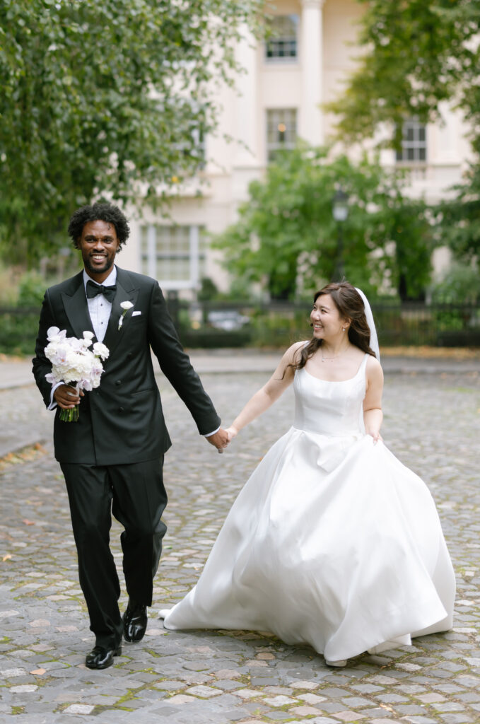 A bride and groom walk hand in hand outdoors on a cobblestone path. The groom wears a black tuxedo and holds a bouquet. The bride wears a white gown and veil. Both are smiling and looking at each other, with greenery in the background.