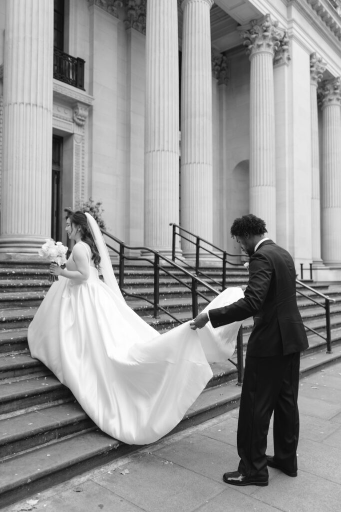 A bride in a long white gown walks up stone steps outside a grand building with columns, while a groom in a suit holds her train. The scene is elegant and formal, captured in black and white.