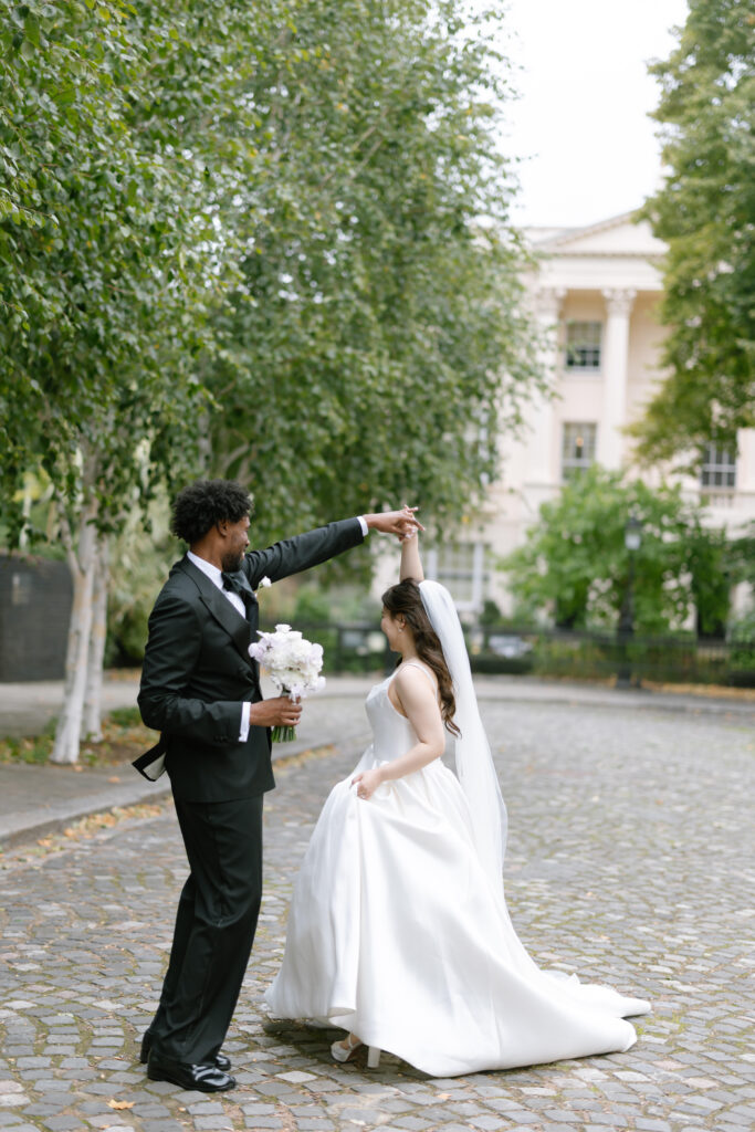 A groom in a black tuxedo twirls his bride, who wears a white wedding dress and veil, on a cobblestone path lined with trees, with a large white building in the background.