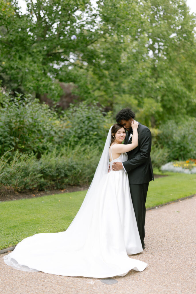 A bride in a white gown and veil embraces a groom in a black suit on a garden path, surrounded by lush green trees and bushes. The bride smiles at the camera while the groom gently kisses her forehead.