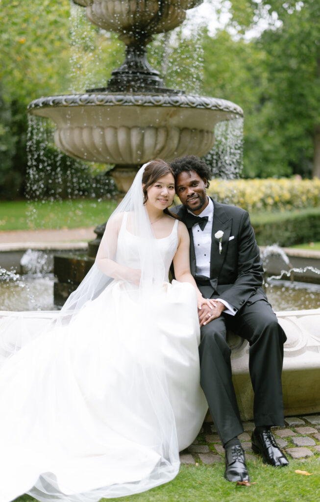 A bride in a white gown and veil sits beside a groom in a black tuxedo on a stone bench in front of a large fountain, both smiling and holding hands in a lush garden setting.