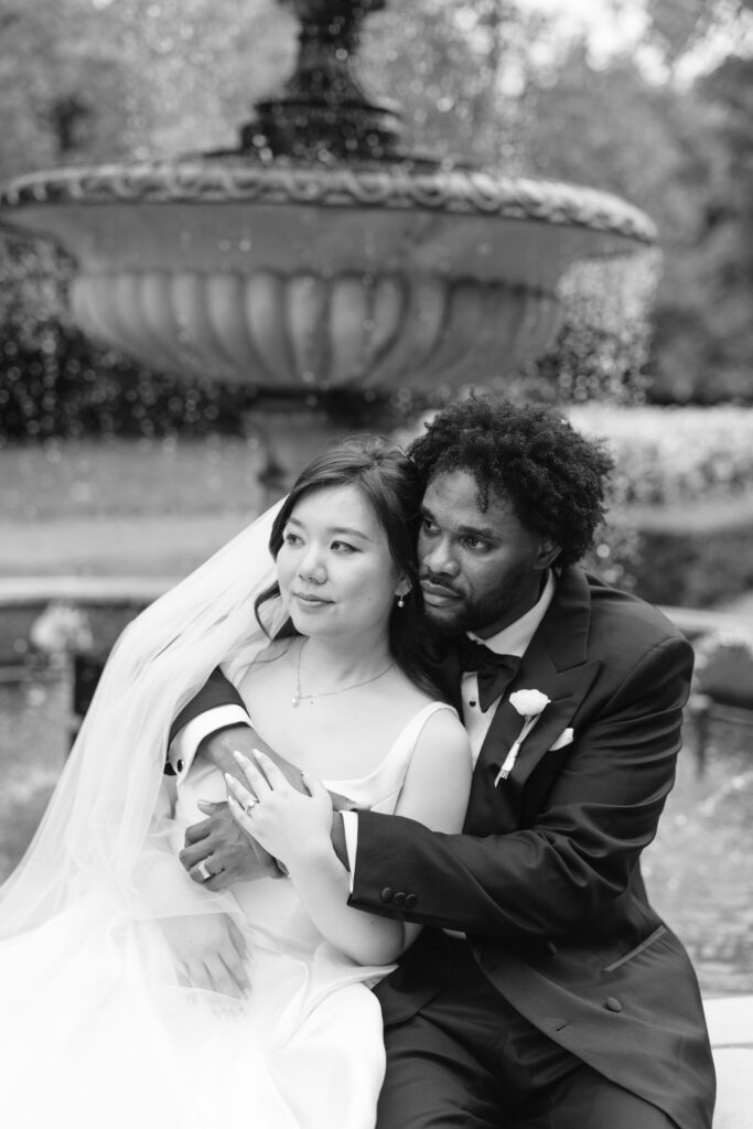 A bride and groom sit closely together in front of a fountain. The bride wears a veil and a sleeveless gown, while the groom, in a tuxedo, gently embraces her. Both appear thoughtful and serene.