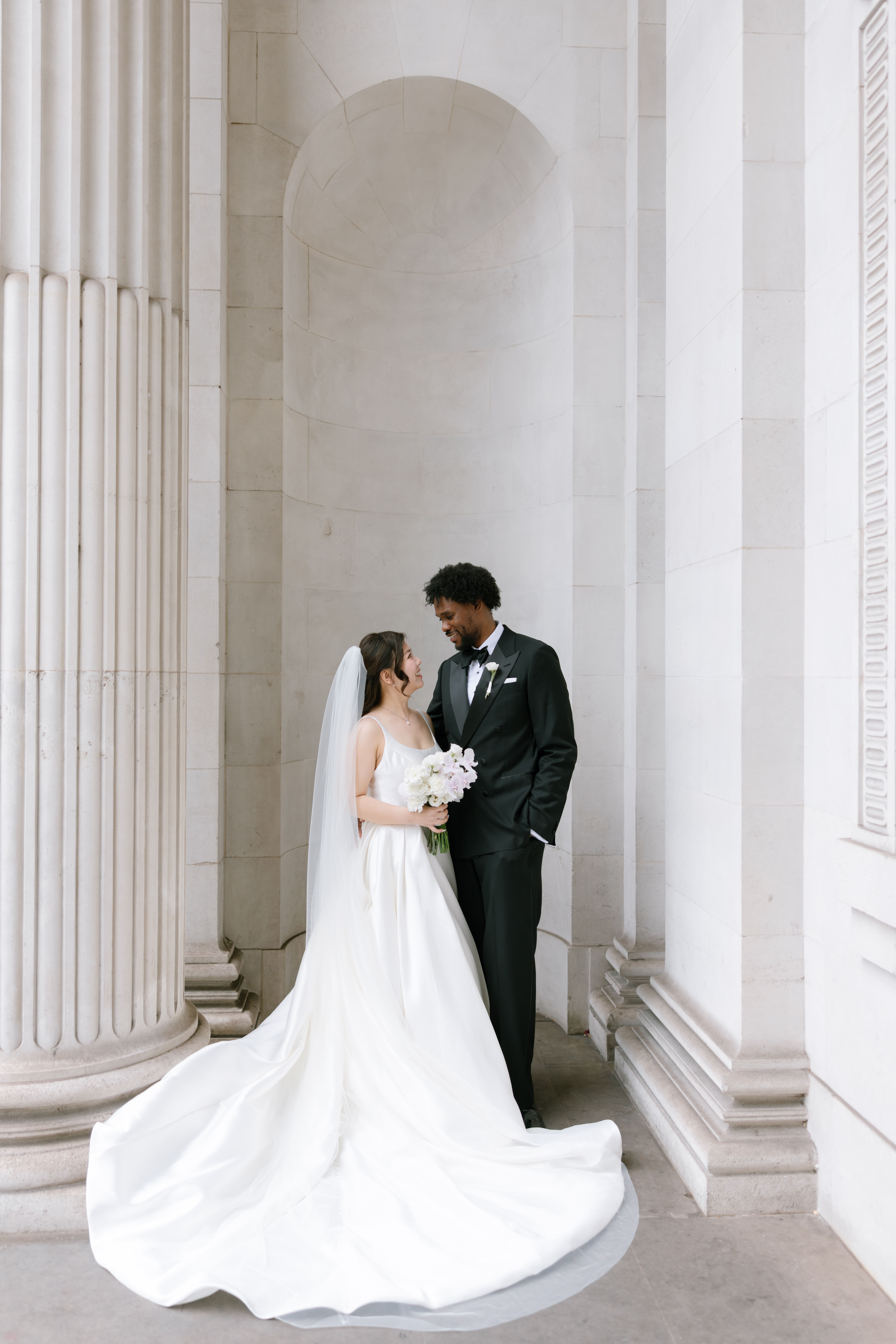 A bride in a white gown and veil holds a bouquet while standing beside a groom in a black suit. They gaze at each other lovingly, surrounded by grand white stone columns and arches.