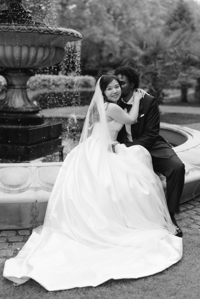 A bride in a flowing white gown and veil embraces a groom in a suit as they sit together on the edge of a fountain in a garden, sharing an intimate moment. Black and white photograph.
