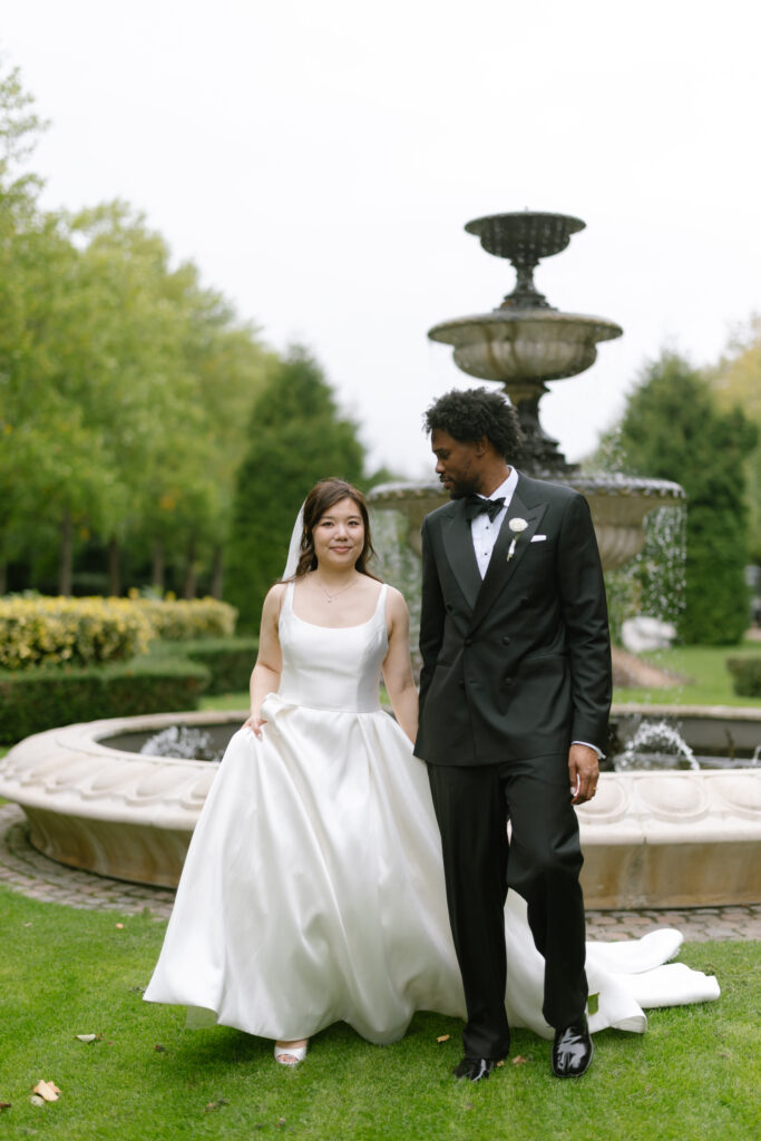 A bride in a white gown and a groom in a black tuxedo walk hand in hand in front of a large stone fountain in a garden setting.