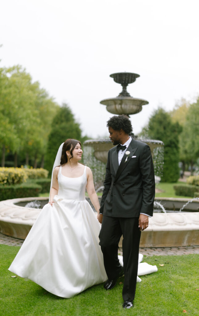 A bride in a white gown and veil holds hands with a groom in a black tuxedo as they walk together on grass in front of a large fountain in a garden. Both are smiling and looking at each other.