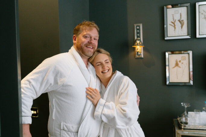 A smiling couple wearing white bathrobes stands close together in a cozy, dark-walled room with framed botanical prints and a small light fixture on the wall behind them.