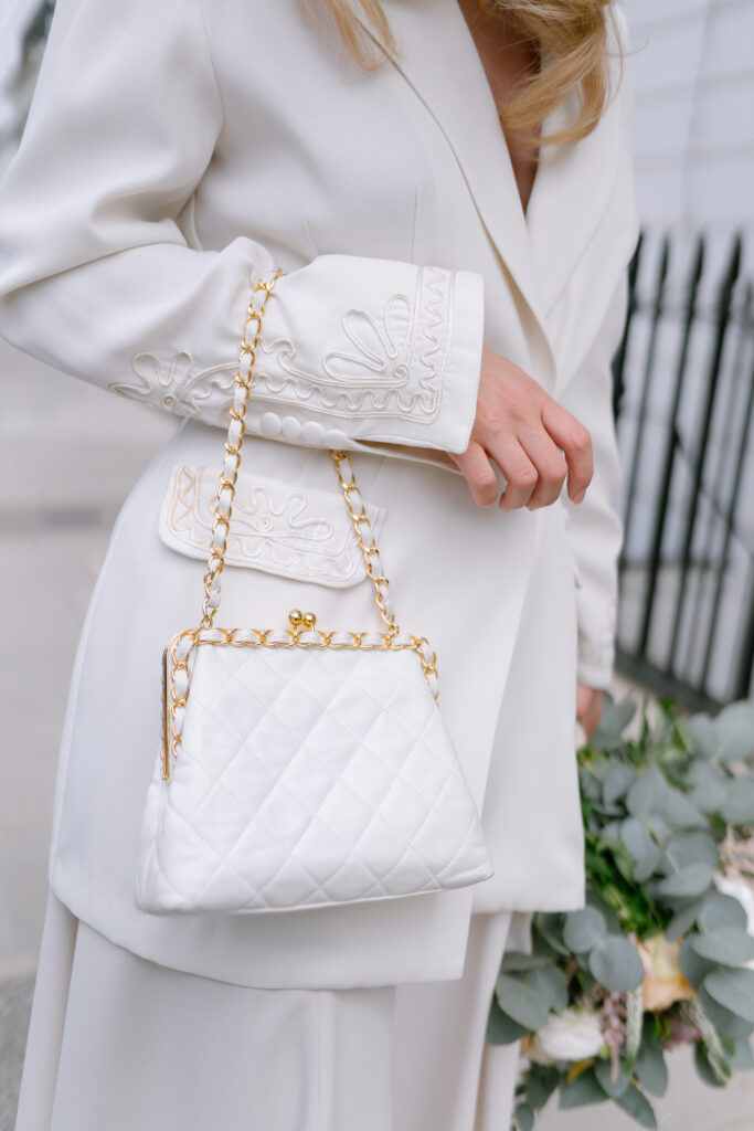 A woman in an elegant white suit with embroidered details holds a white quilted handbag with a gold chain strap. She also carries a bouquet of greenery and white flowers. The background shows a blurred outdoor setting.