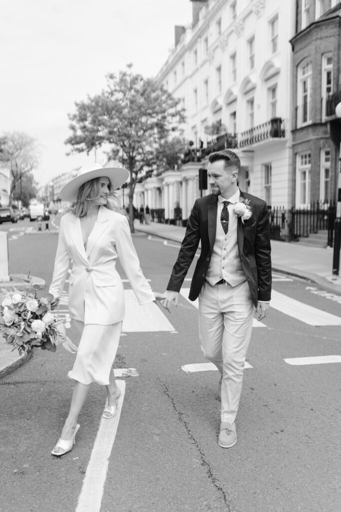 A bride and groom walk hand in hand across a city street. The bride wears a white dress, blazer, and wide-brimmed hat, holding a bouquet, while the groom wears a light suit with a dark jacket. The background shows townhouse buildings.