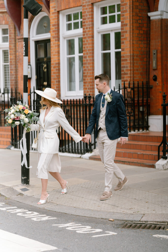A couple dressed in elegant attire holds hands while crossing a city street. The woman wears a white suit, wide-brimmed hat, and heels, while the man wears a navy blazer and beige trousers. Both have boutonnières.