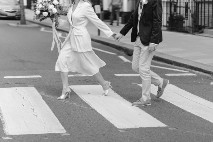 A couple dressed in wedding attire holding hands and crossing a street on a crosswalk, with the woman carrying a bouquet. The image is in black and white.