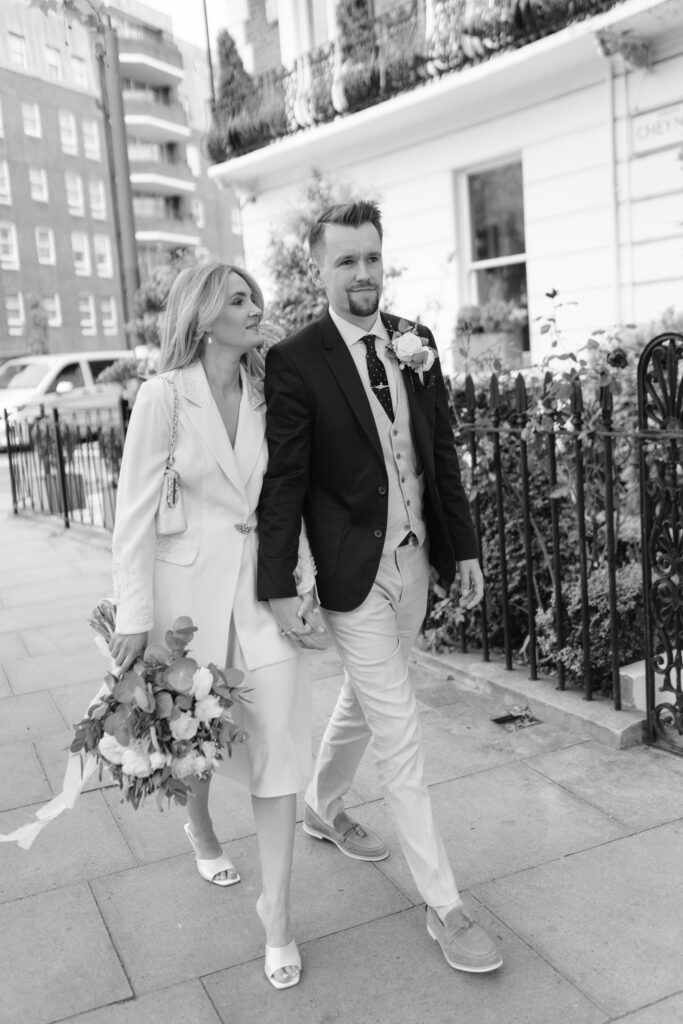 A couple dressed in wedding attire walk hand in hand on a city sidewalk. The woman wears a white blazer-style dress and holds a bouquet, while the man wears a suit with a boutonnière. The photo is in black and white.