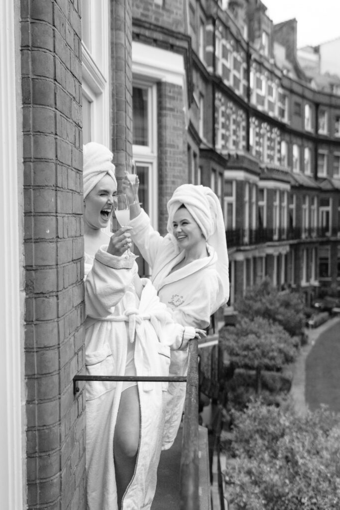 Two women in bathrobes and towels on their heads laugh and toast with drinks while leaning out of a window on an upper floor of a brick building, with a row of similar buildings visible in the background.