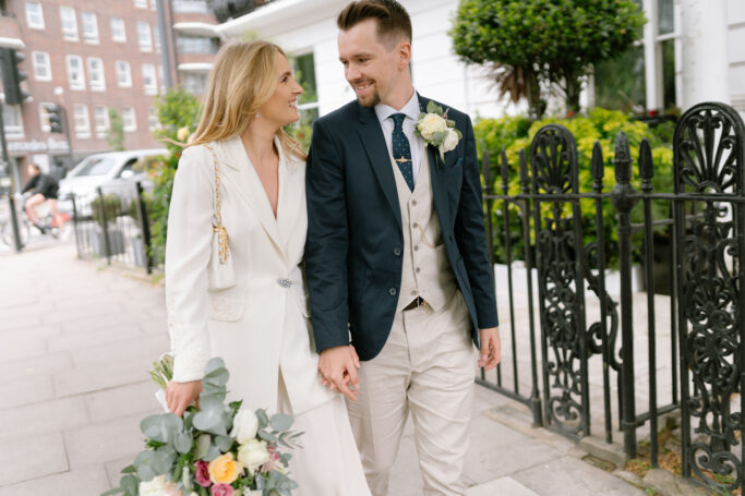 A smiling couple dressed in wedding attire holds hands while walking down a city sidewalk. The woman wears a white dress and carries a bouquet; the man wears a suit with a vest and boutonniere. They look at each other fondly.