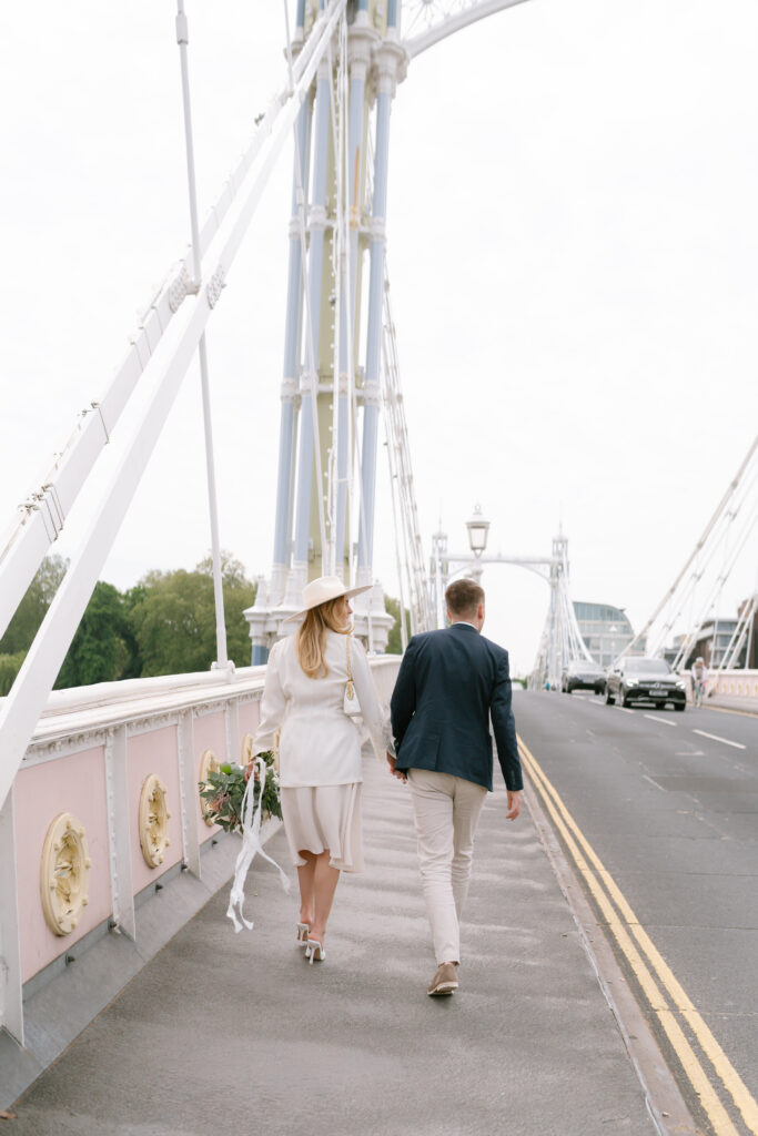 A couple, dressed in light, semi-formal outfits, walk hand-in-hand across a decorative suspension bridge. The woman holds a bouquet and wears a wide-brimmed hat. The scene is bright with soft daylight.