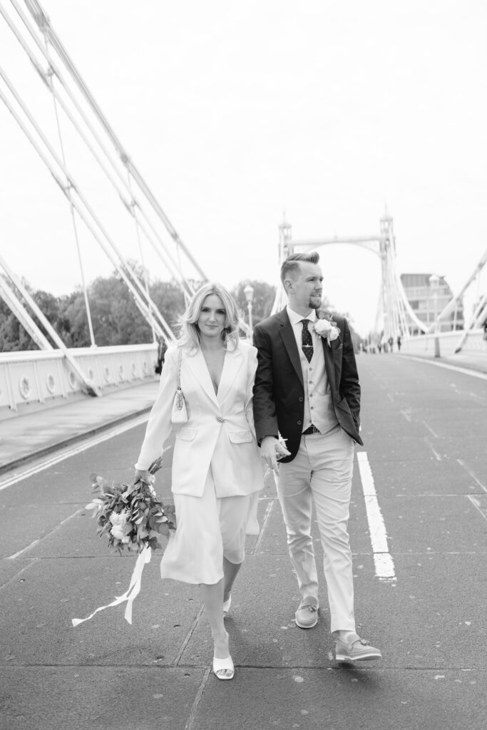 A couple dressed in wedding attire walks hand in hand down a bridge. The woman holds a bouquet and wears a white suit, while the man wears a suit with a boutonniere. The scene is in black and white.