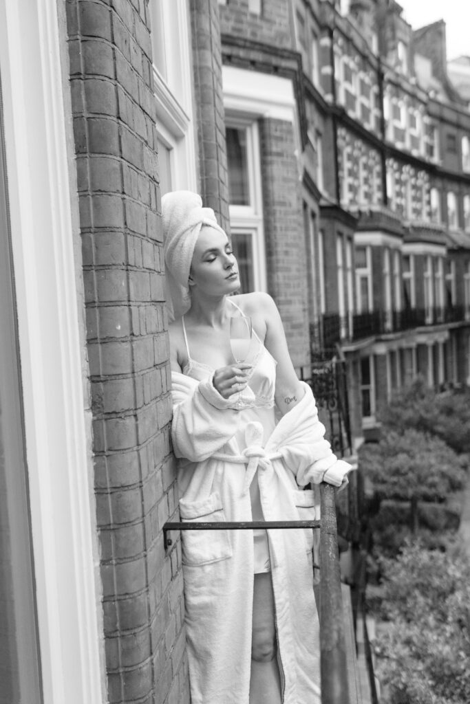 A person in a bathrobe and towel stands on a balcony, holding a glass and looking relaxed. The black-and-white image shows brick buildings and green trees in the background.