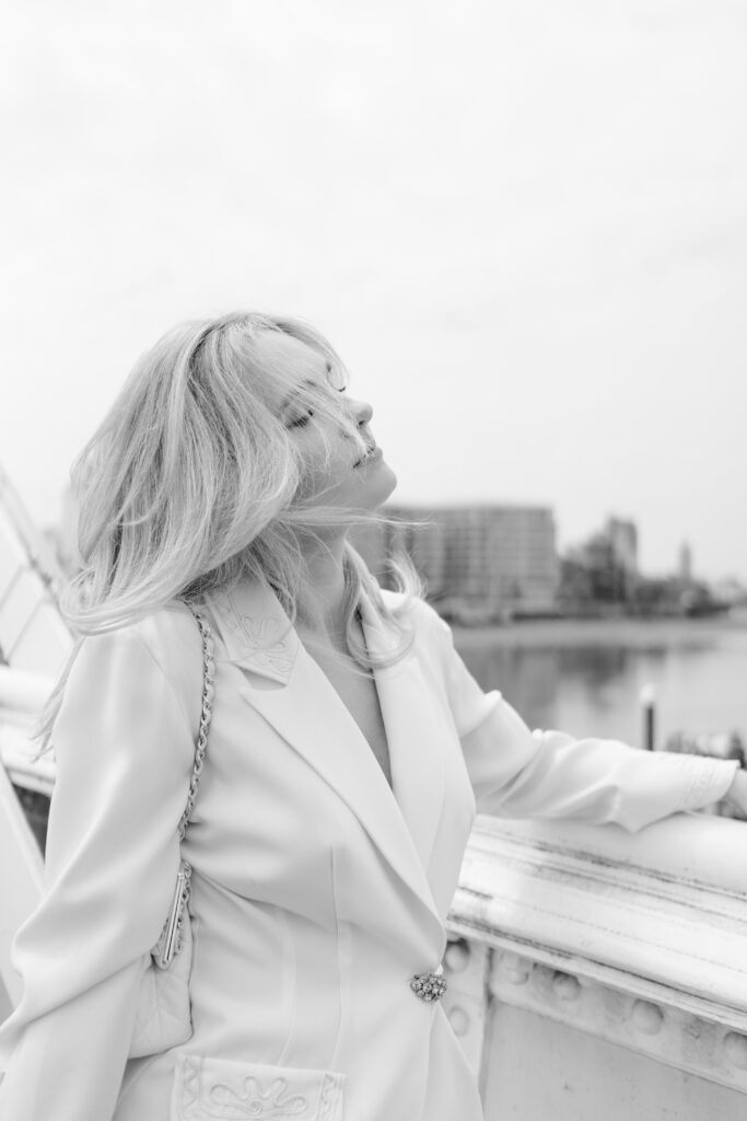 A woman with light hair wearing a white blazer stands outdoors by a railing, looking upward with her eyes closed. The background shows a blurred cityscape and water. The image is in black and white.