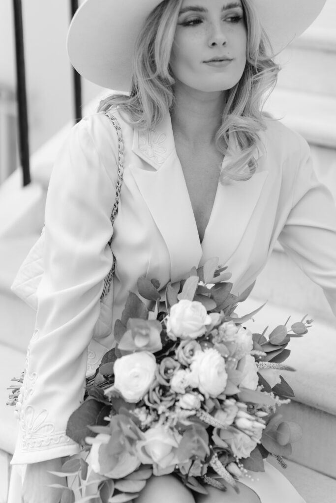 A woman in a white hat and suit sits on steps, holding a large bouquet of flowers. The image is black and white, highlighting her light hair and elegant attire.