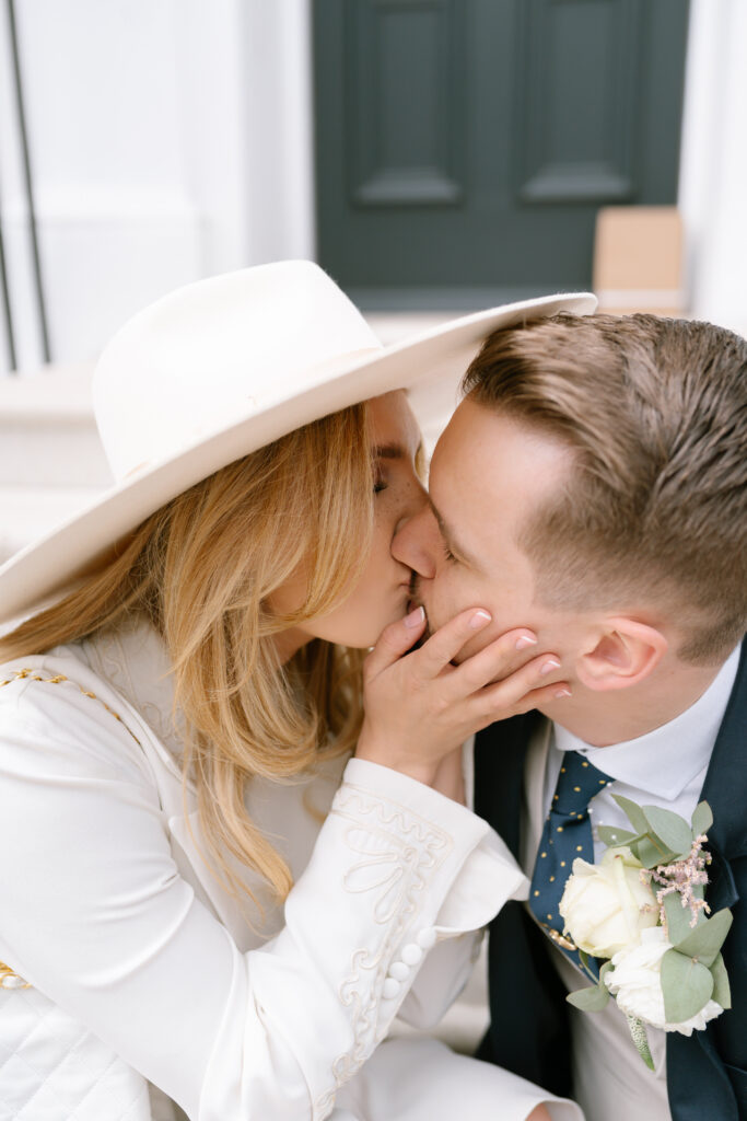 A couple dressed in wedding attire share a kiss. The woman, wearing a white hat and embroidered jacket, gently holds the mans face. The man is in a suit with a boutonnière pinned to his lapel.
