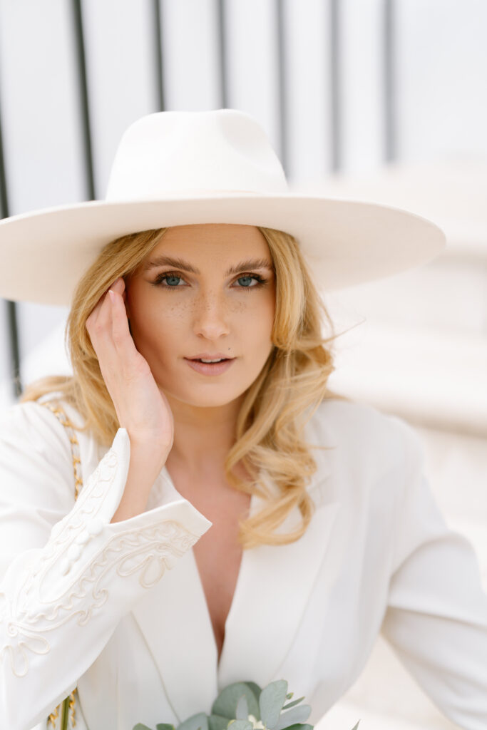 A woman with blonde hair wearing a wide-brimmed white hat and a white embroidered jacket looks at the camera, gently touching the side of her face. She is sitting on white steps, and holding greenery.