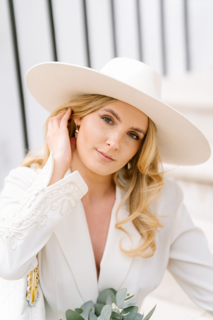 A woman with long blonde hair wears a wide-brimmed white hat and a white outfit with embroidered details. She looks at the camera, gently touching her hair, with a neutral background and some greenery visible.