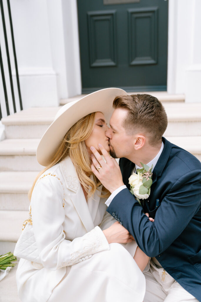 A couple dressed in elegant attire sits on white steps, sharing a kiss. The woman wears a wide-brimmed hat and white suit, holding the mans face. The man wears a navy suit, holding a small bouquet of white roses.