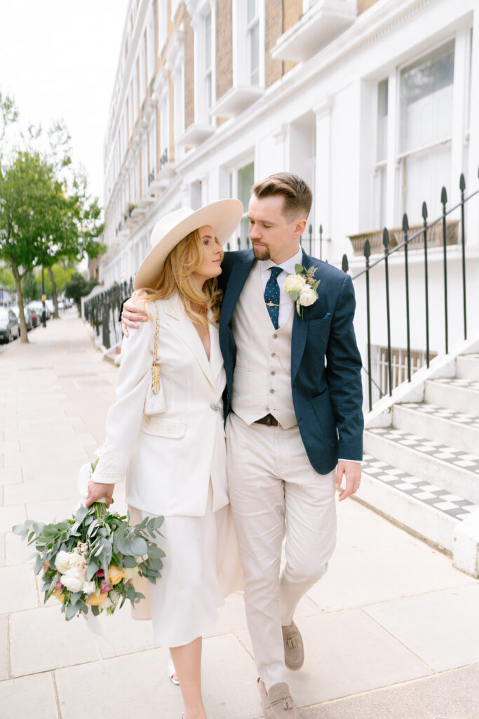A couple walks arm-in-arm on a city sidewalk. The woman wears a white suit, hat, and holds a bouquet; the man wears a blue jacket, light vest, and tie. They look at each other affectionately. Townhouses line the street.