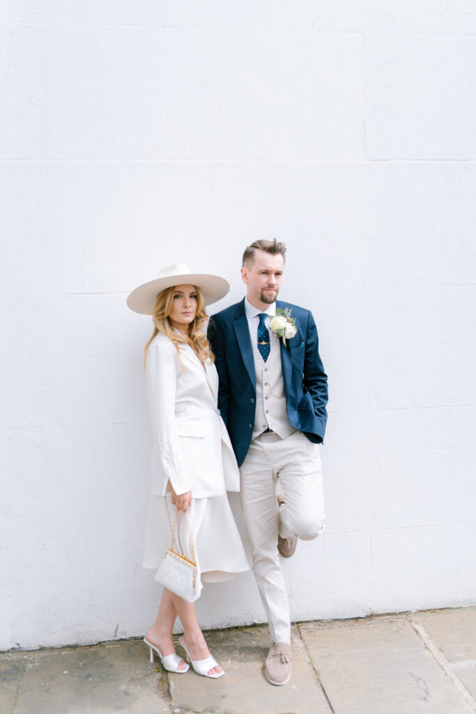 A woman in a white dress, wide-brim hat, and heels stands next to a man in a blue blazer, beige vest, and trousers. They pose against a plain white wall, both looking at the camera with relaxed expressions.