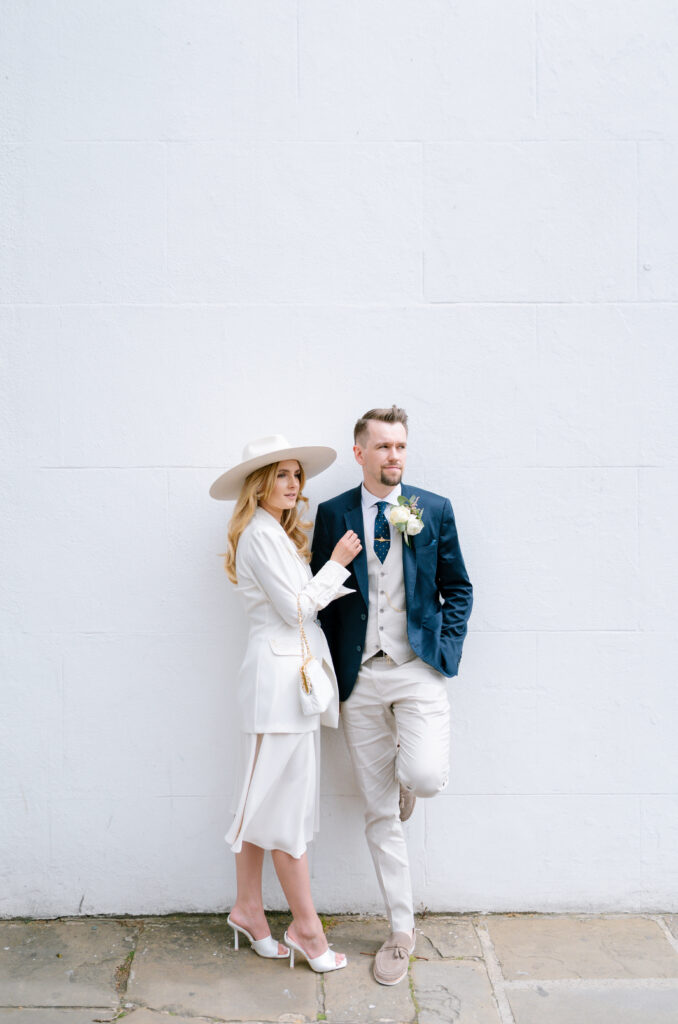 A stylish couple poses against a white wall. The woman wears a white dress, blazer, wide-brimmed hat, and heels. The man wears a navy blazer, light vest, tie, boutonnière, and pale pants, standing with one foot against the wall.