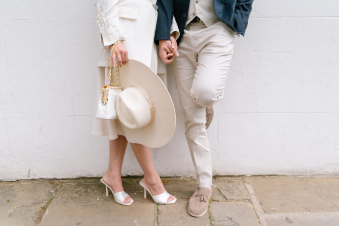 A stylish couple stands against a white wall, holding hands. The woman wears heeled sandals, a white dress, and holds a wide-brim hat and a purse. The man wears light pants, a jacket, and loafers. Neithers face is visible.