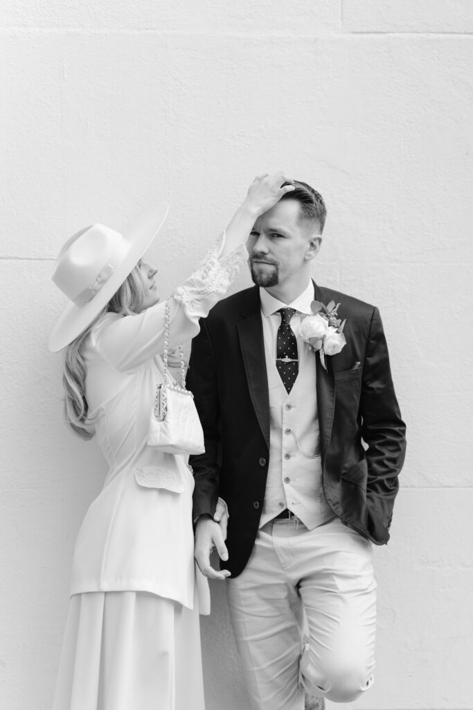 A woman in a wide-brimmed hat playfully touches the hair of a man in a suit and boutonniere. They stand against a plain light-colored wall, and both have relaxed, expressive poses. The image is in black and white.