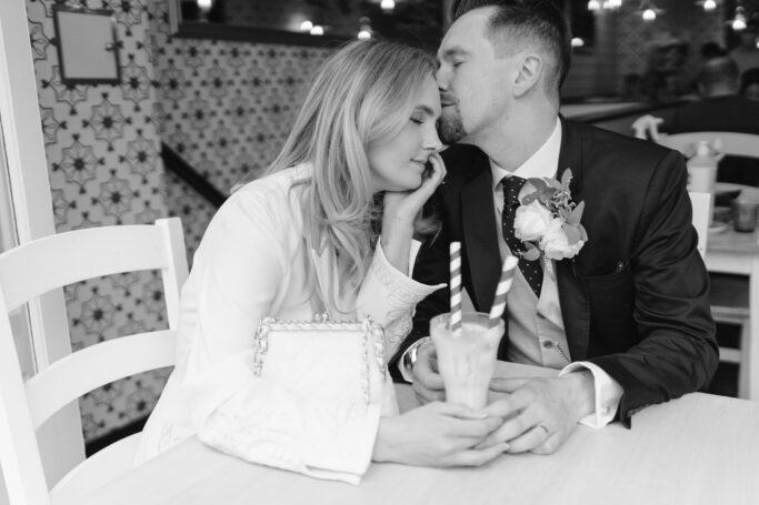 A couple sits closely at a table. The man in a suit gently kisses the woman’s forehead. She smiles with her eyes closed, holding a milkshake with striped straws. The setting has patterned tiles and a warm atmosphere.