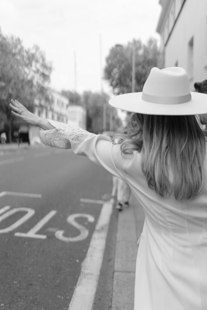 A woman in a white hat and dress stands on the edge of a street with her arm raised, as if hailing a cab. The photo is in black and white and taken from behind. Trees and buildings are visible in the background.