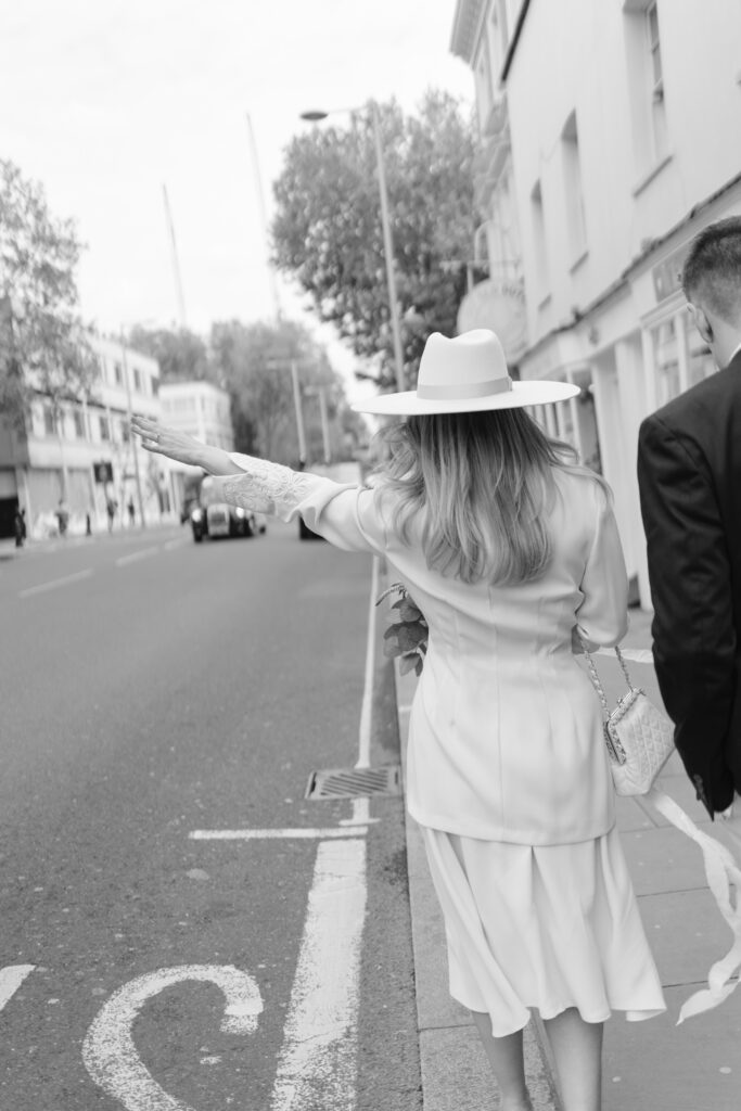 A woman in a light dress and wide-brimmed hat stands on a city sidewalk, facing away and extending her arm as if to hail a taxi. A man in a suit walks beside her. The image is black and white.
