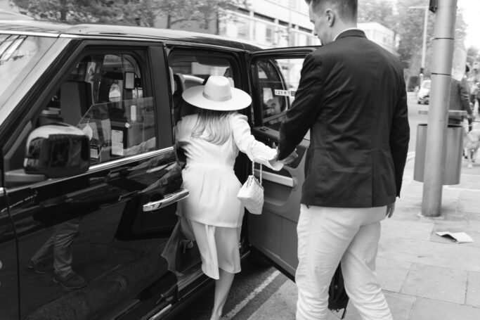 A woman in a white outfit and wide-brimmed hat steps into a car while a man in a jacket and light pants holds her hand and the car door open, on a city street.