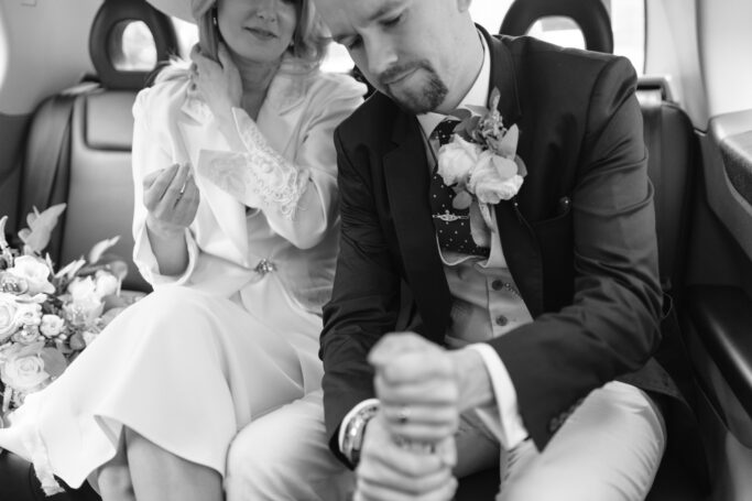 A bride and groom sit in the back seat of a car. The bride adjusts her hat and holds flowers, while the groom, wearing a suit with a boutonniere, focuses on opening a bottle. The image is in black and white.
