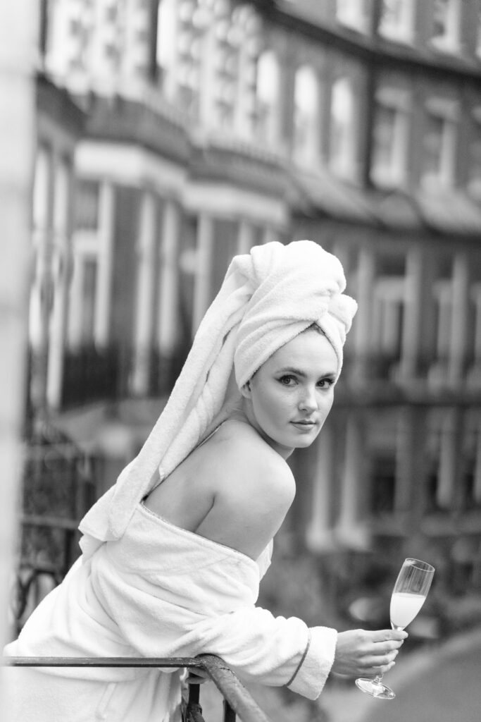 A woman in a bathrobe and towel turban leans on a balcony railing, holding a glass of champagne, with blurred city buildings in the background. The image is in black and white.