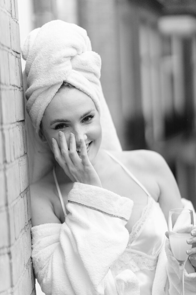 A woman in a bathrobe and towel turban stands by a brick wall, smiling with one hand to her face and holding a glass of milk in the other. The image is in black and white.