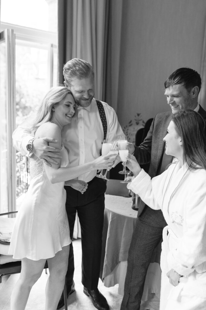 Four people dressed formally stand together indoors, smiling and toasting with drinks. The scene appears joyful and celebratory, with natural light coming from an open window in the background. The image is in black and white.