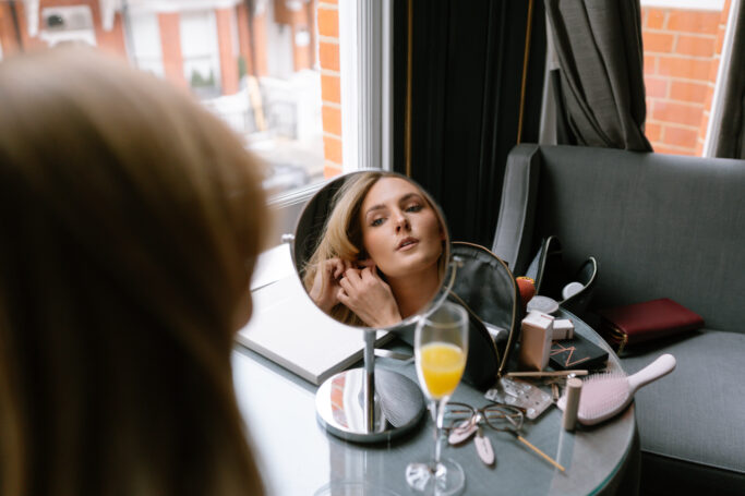 A woman sits at a table by a window, looking into a round mirror as she puts on an earring. On the table are a drink, glasses, makeup items, a notebook, and a hairbrush.