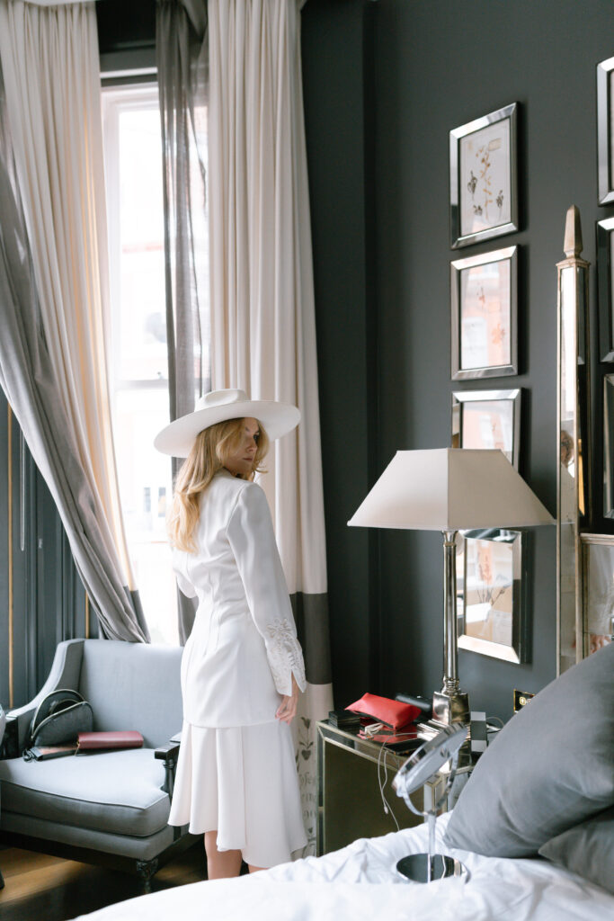 A woman in a white outfit and wide-brimmed hat stands in a stylish, modern room with dark walls, framed art, a gray chair, and a large window with sheer curtains. She looks over her shoulder toward the camera.
