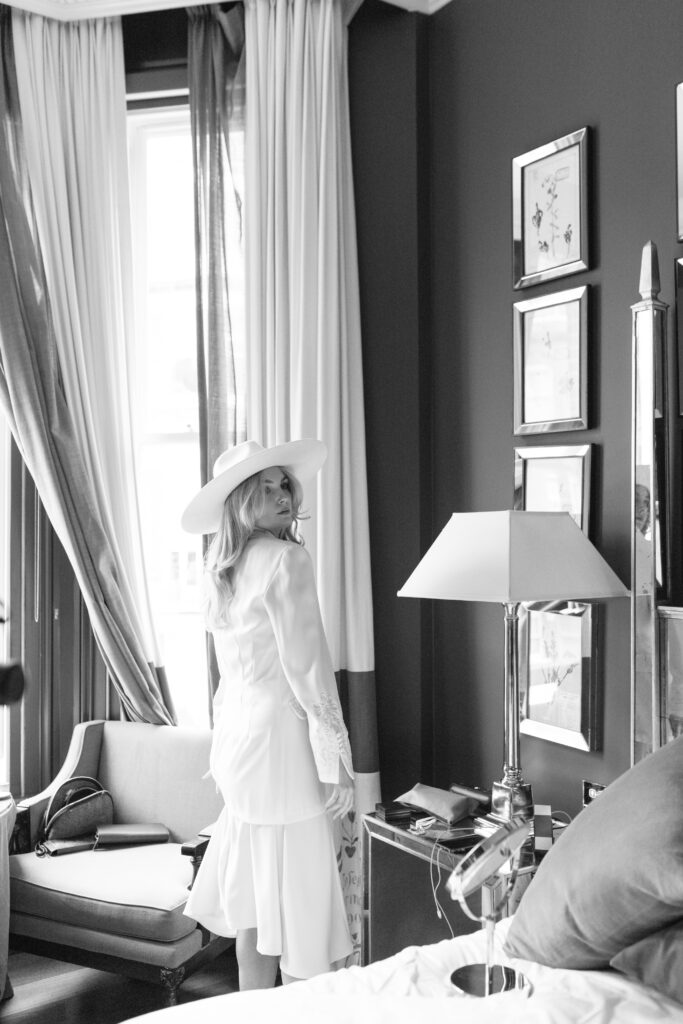 A woman in a white dress and wide-brimmed hat stands in a stylish, softly lit room near a window, glancing back with a gentle smile. The room features framed art, a lamp, and elegant furnishings.