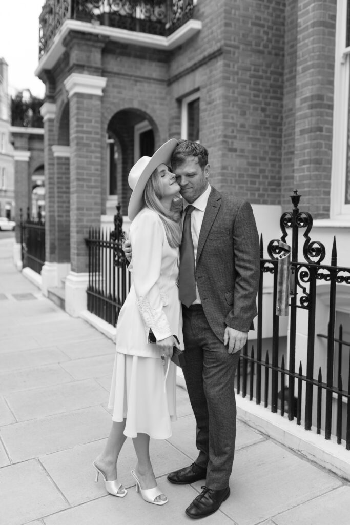 A woman in a white dress and wide-brimmed hat kisses a man in a suit on the cheek. They stand close together on a city sidewalk, with brick buildings and wrought iron railings in the background.