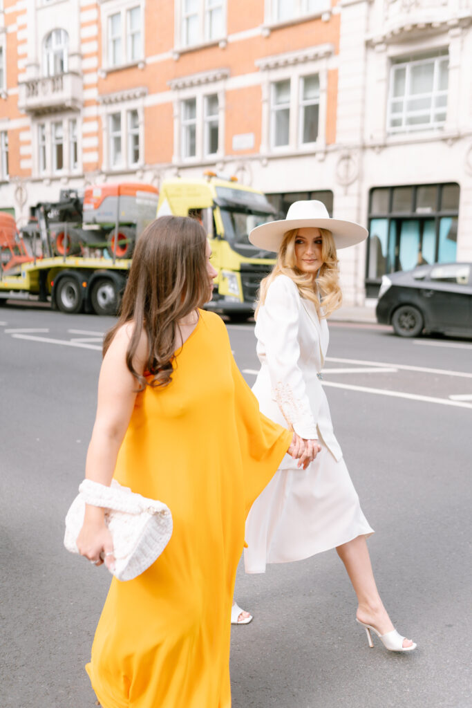 Two women cross a city street. One wears a bright yellow dress and holds a white clutch, the other wears a white dress, white hat, and heels. They hold hands and smile, with buildings and vehicles in the background.