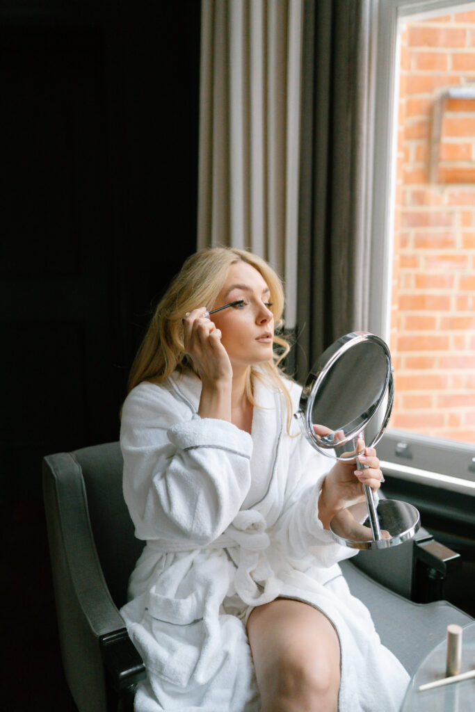A woman in a white bathrobe sits by a window, holding a handheld mirror and applying makeup to her face. Natural light comes through the window, illuminating her as she concentrates on her reflection.