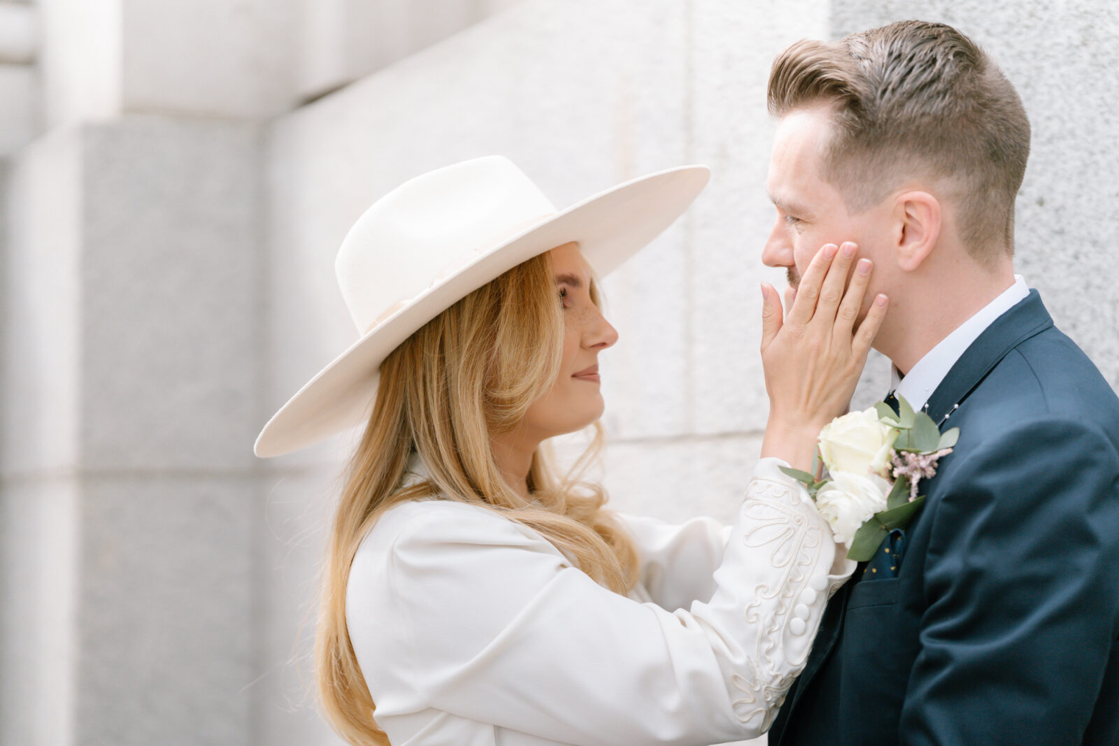 A woman in a white hat gently touches the face of a man in a dark suit with a white rose boutonniere. They stand close together, smiling softly, in front of a light stone wall.