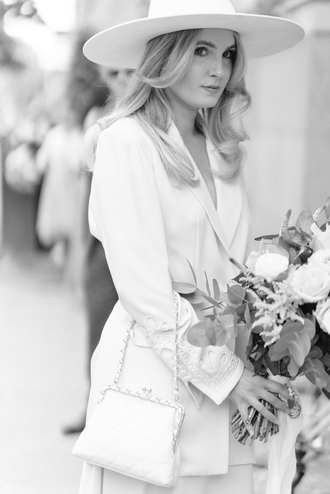 A woman in a wide-brimmed hat and elegant light-colored outfit holds a bouquet of flowers and a quilted handbag, looking at the camera. The photo is in black and white.
